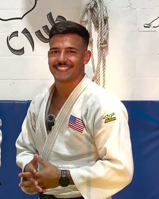 Sensei Alejandro Menendez smiling in his judogi in front of the dojo mural.