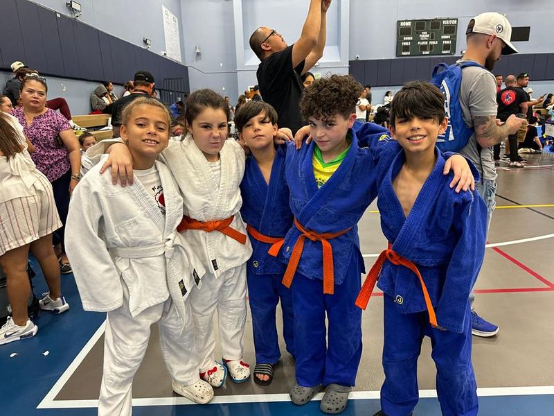 Five kids in judogis with orange belts smiling together at a tournament.