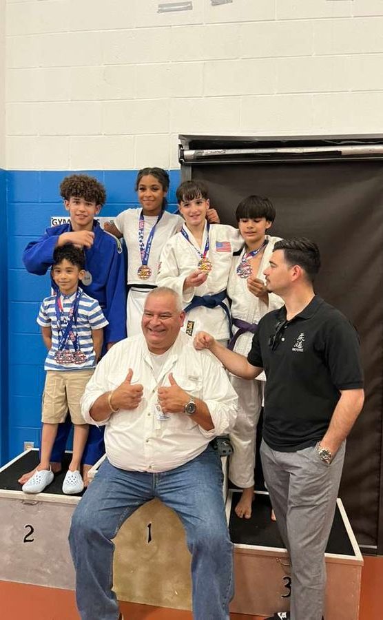 A coach giving two thumbs up on a podium, with young students wearing their medals behind him.