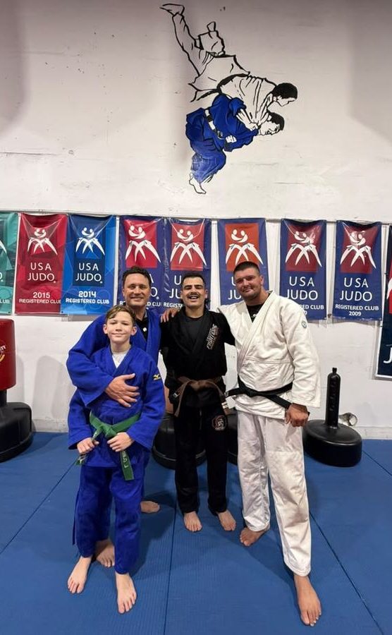 A young student with a green belt standing with three coaches under the USA Judo banners.