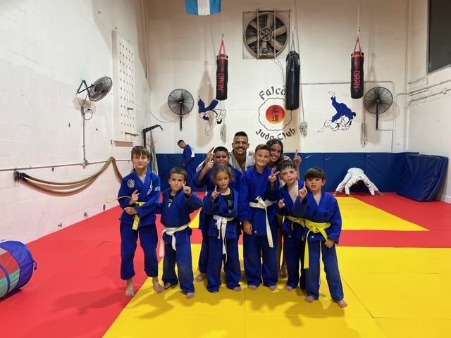 A group of kids in blue judogis smiling in front of the Falcon Judo Club mural.