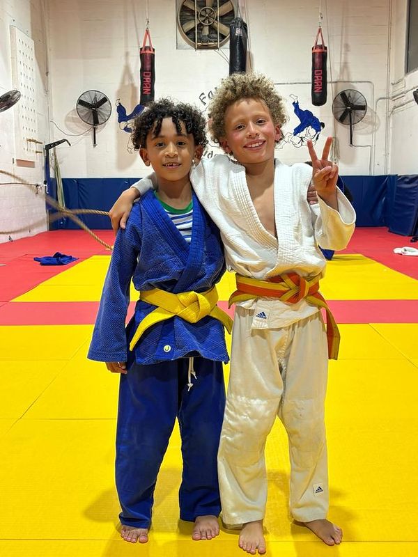 Two young students hugging on the red and yellow mat, one in a blue judogi and one in white.